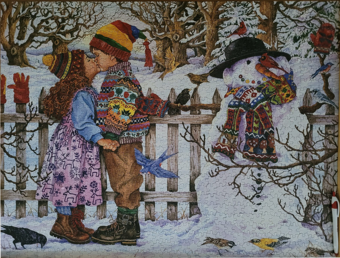 A young boy and girl kissing in the snow next to a snowman in a winter garden.