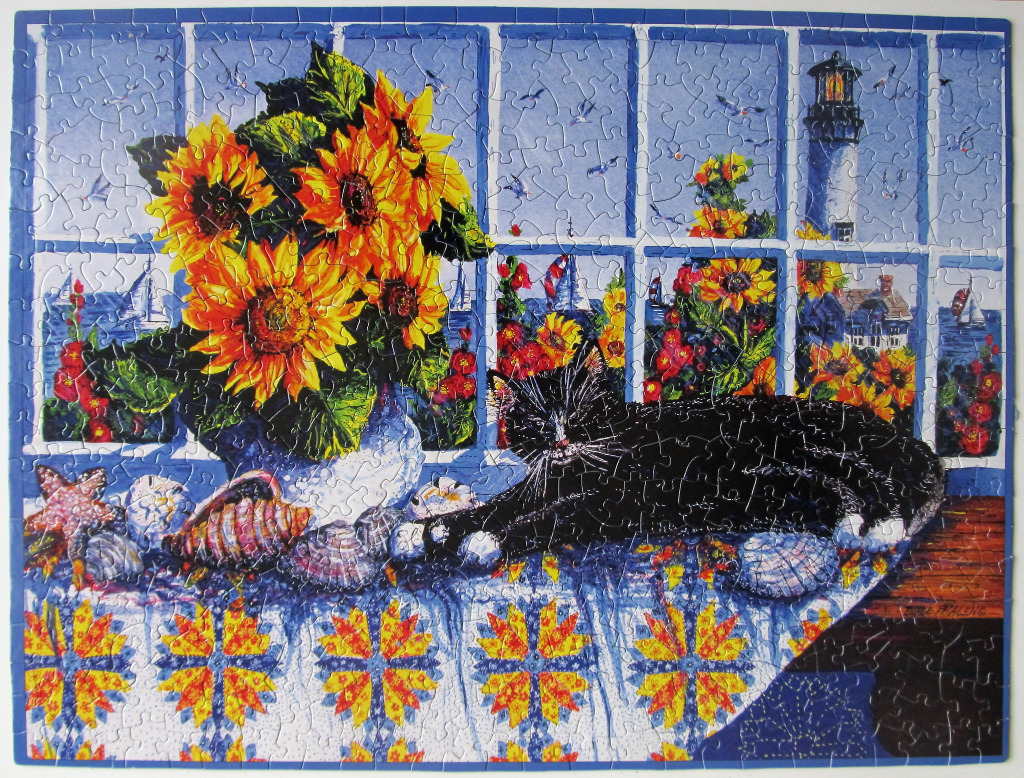 A black and white cat resting on a quilt near a window with sunflowers and a seaside view.