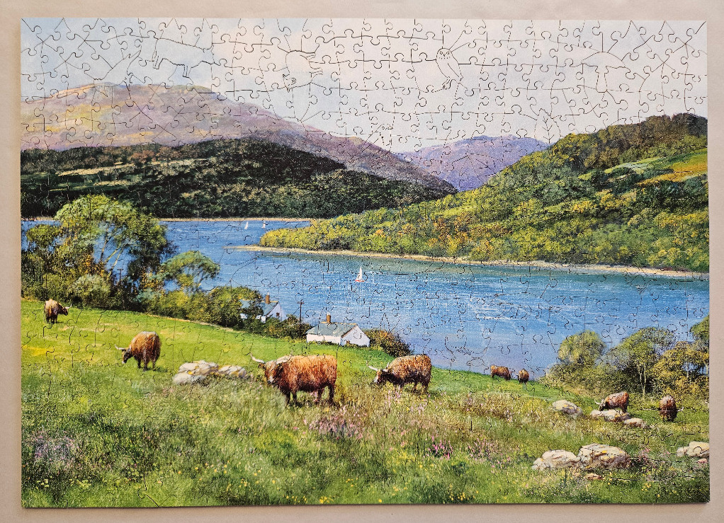 A Scottish Highland landscape featuring cattle grazing in a meadow beside a blue lake and rolling hills.