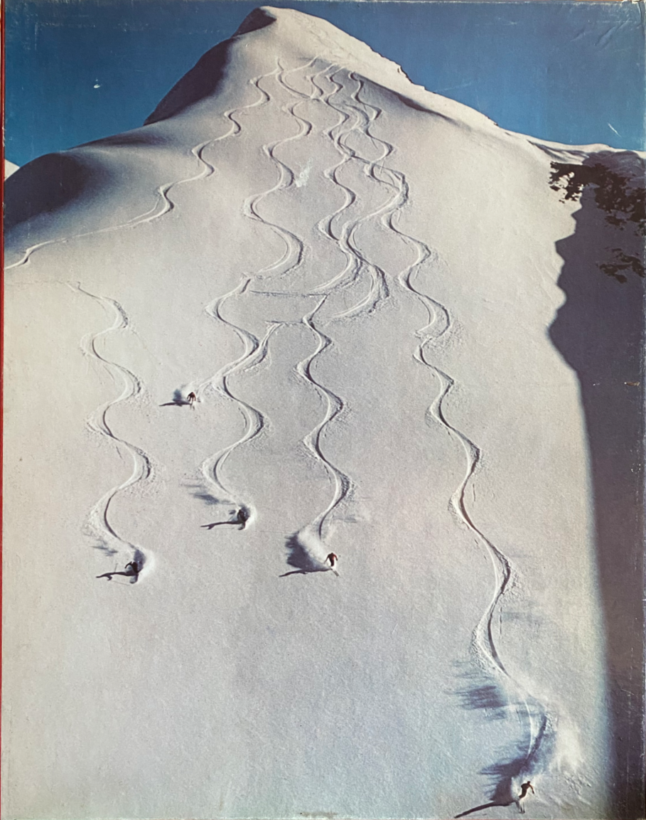 Skiers descending a vast, sunlit, snow-covered mountain slope with intricate winding tracks behind them.