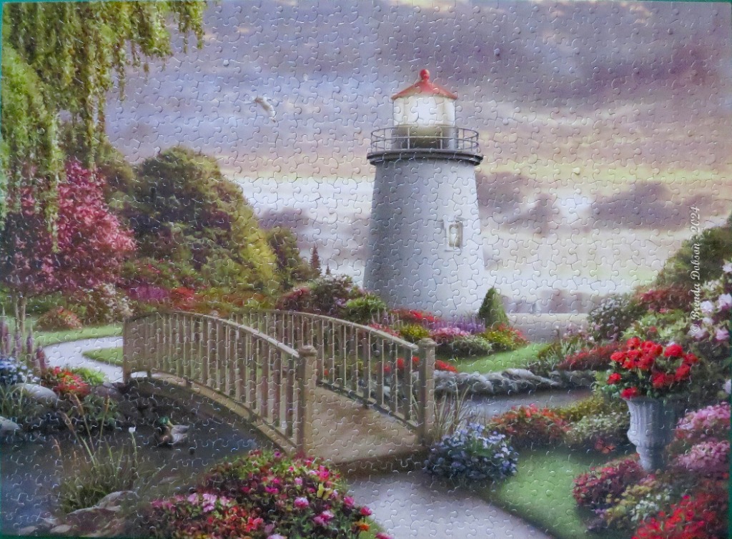 A lighthouse illuminated at twilight in a vibrant, flower-filled coastal garden with a wooden footbridge over a stream.
