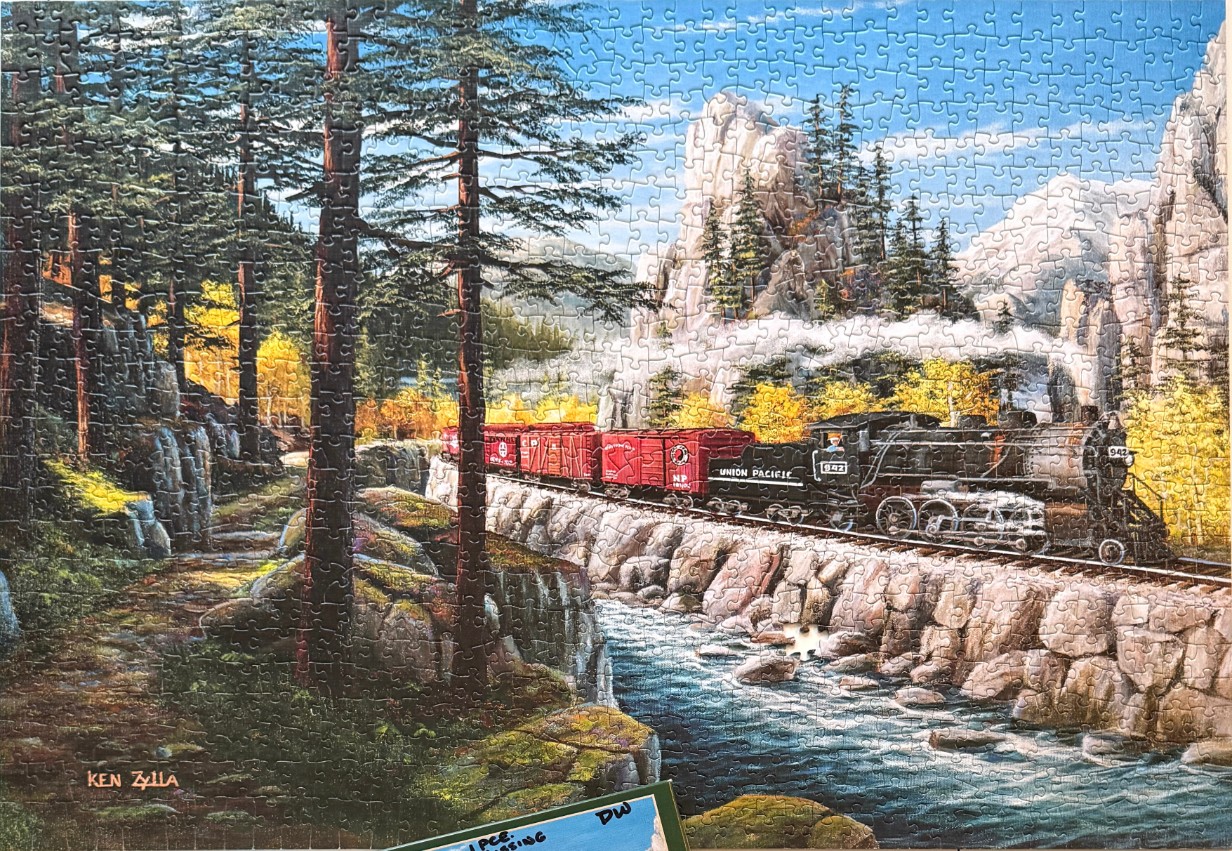 A steam train traveling along a track next to a mountain stream surrounded by evergreen trees and rocky cliffs.