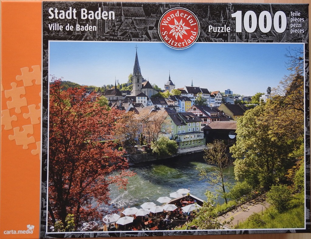 A sunny, scenic view of the Swiss town of Baden with a river, historic architecture, and lush trees.