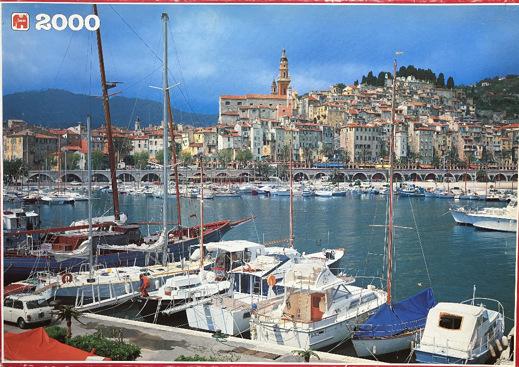 A sunny, detailed photograph of the harbor in Menton, France, featuring boats in the foreground and a hillside city.