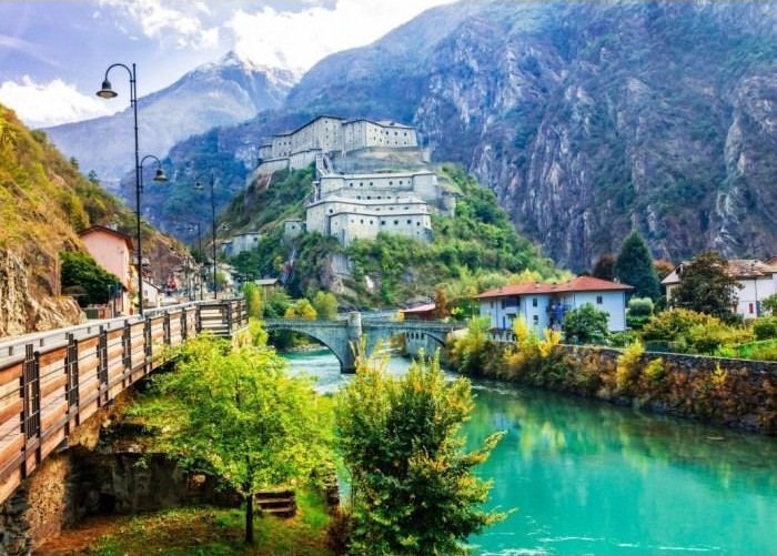 A stone fortress perched on a hill above a turquoise river and a stone bridge, set against high mountains.