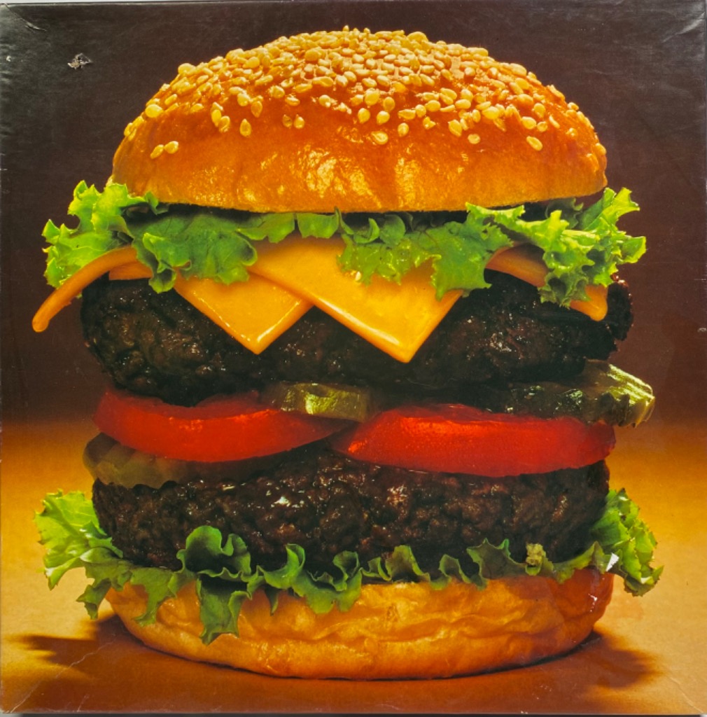 A close-up photograph of a double cheeseburger with lettuce, tomato, and pickles centered against a dark brown background.