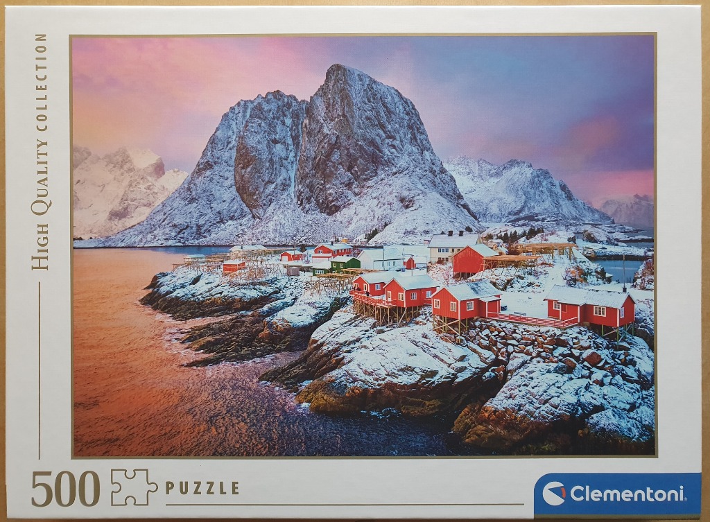 A small coastal village of red houses nestled at the base of a snowy mountain peak under a pink and purple sunset.