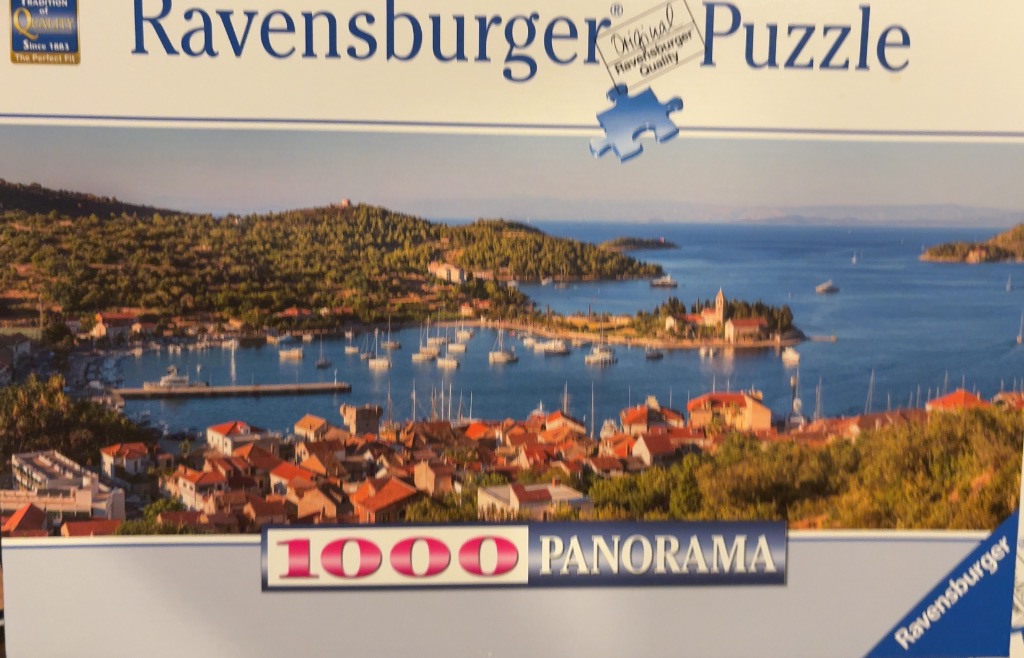 A panoramic coastal view of a Mediterranean village with red-tiled roofs nestled along a harbor full of sailboats.