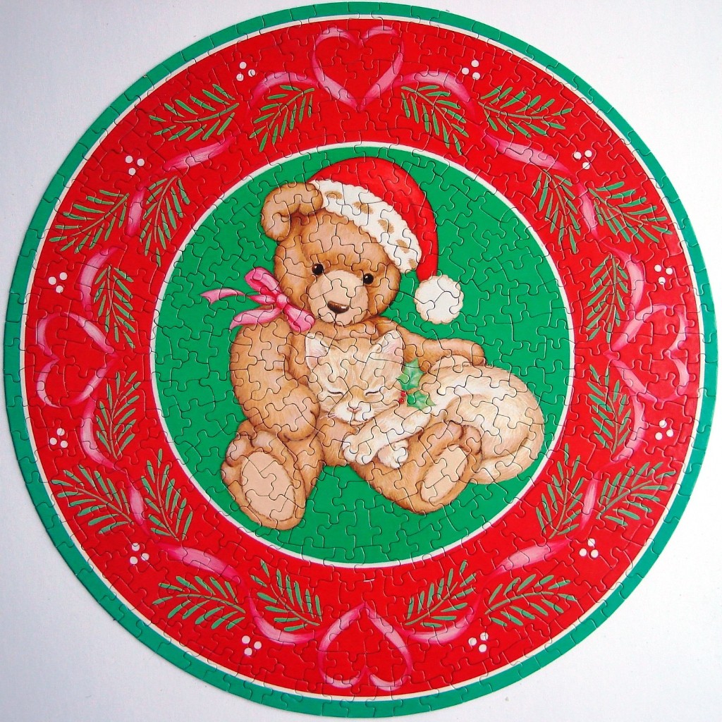 A round illustration of a teddy bear wearing a Santa hat cuddling a kitten, surrounded by a red and green wreath border.