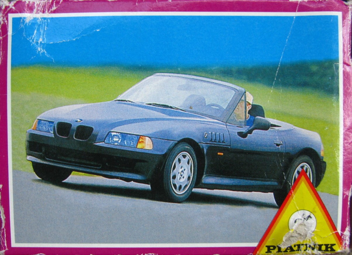 A blue BMW Z3 roadster driving on an asphalt road against a blurred background of green fields and a blue sky.