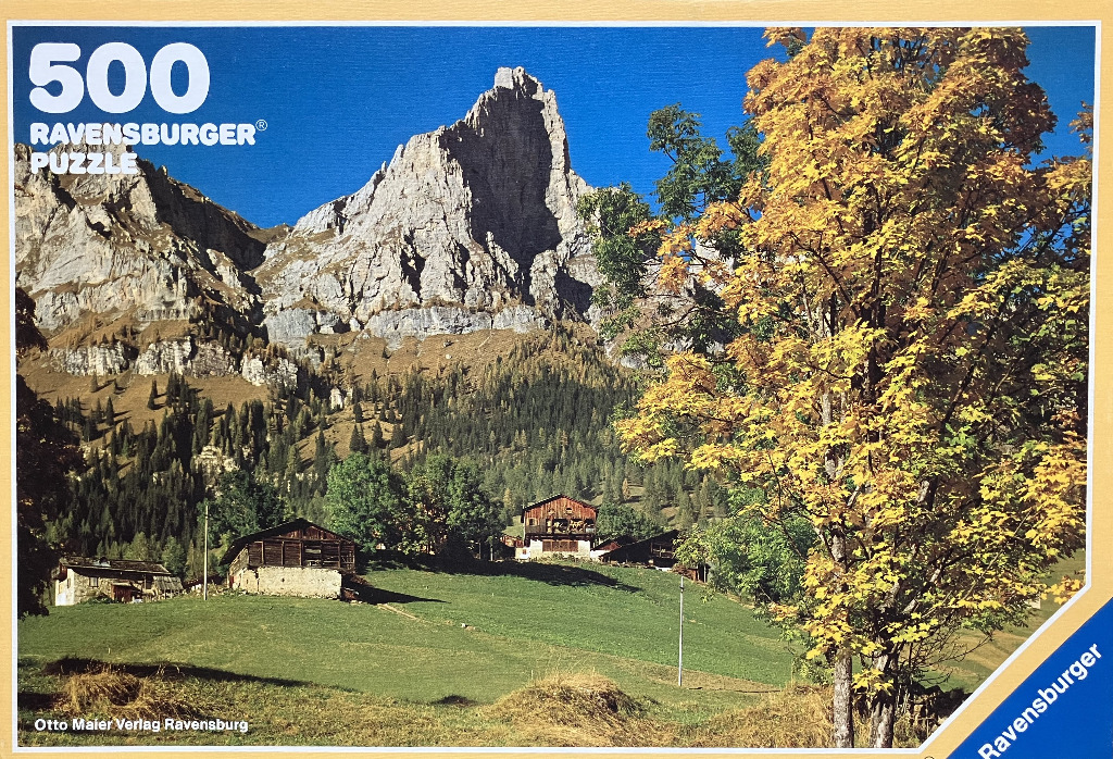 A photograph of a rugged Alpine mountain peak rising behind a green meadow with wooden cabins and an autumn tree.