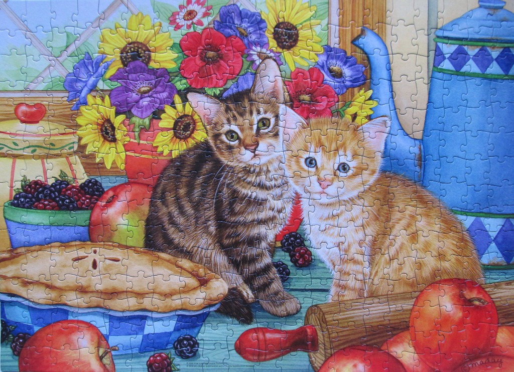 Two kittens sitting on a kitchen counter with flowers, fruit, and a pie.