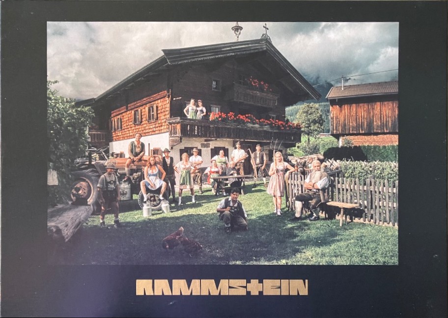 A group of people dressed in traditional Bavarian folk attire posed in front of a rustic wooden farmhouse.
