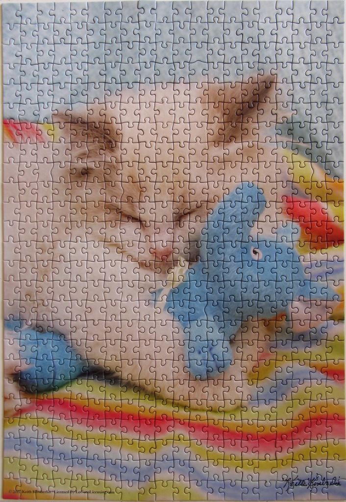 A fluffy cream-colored kitten sleeping while cuddling a blue stuffed toy on a rainbow-striped blanket.