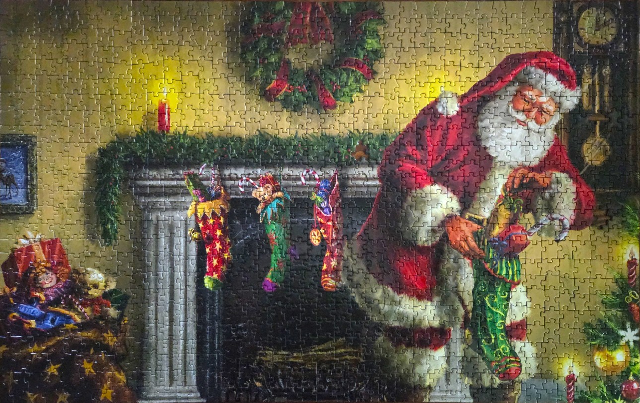 Santa Claus filling a stocking by a fireplace with Christmas decorations