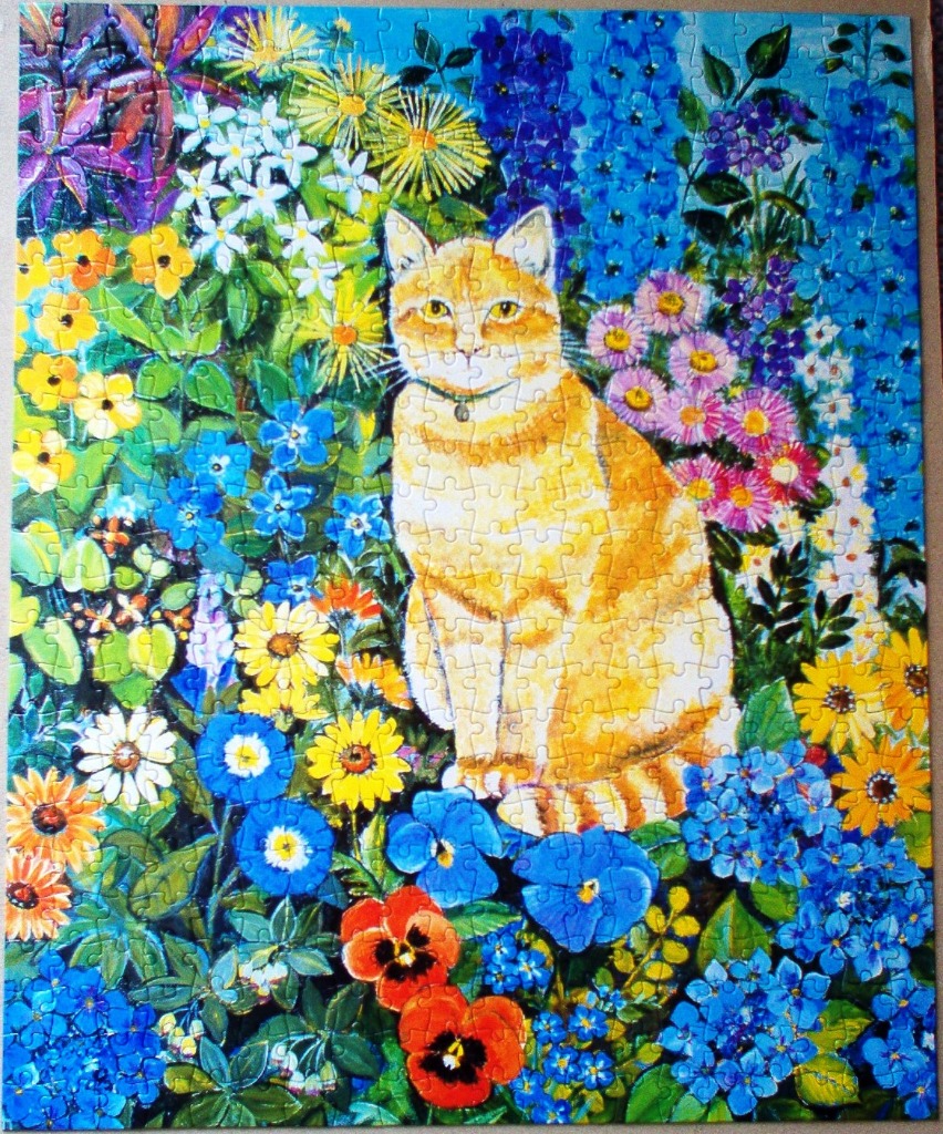 An orange tabby cat sitting in a lush, vibrant garden filled with bright blue, yellow, and multi-coloured flowers.