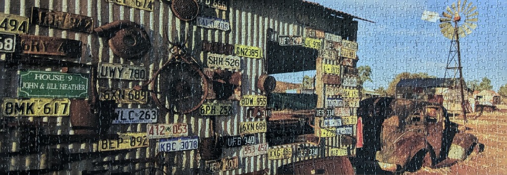 A wall covered in vintage license plates next to a rusted car and a windmill in a desert setting.