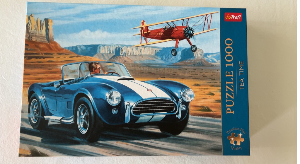 A blue vintage sports car driving through a desert landscape with a red biplane flying overhead.