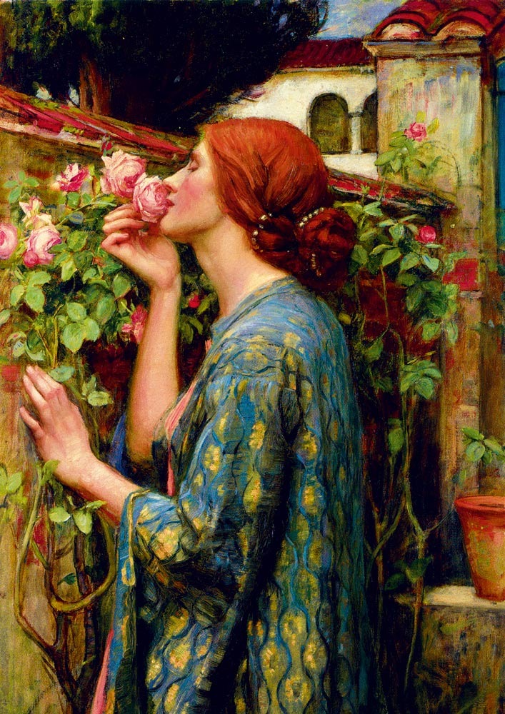 A woman with auburn hair in a blue patterned robe smelling a pink rose by a stone wall.