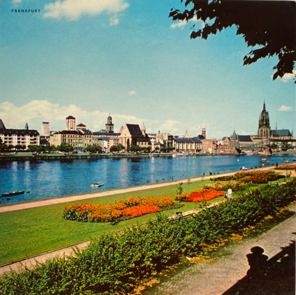 View of the Frankfurt skyline across the Main river with a foreground park featuring vibrant flower beds and hedges.