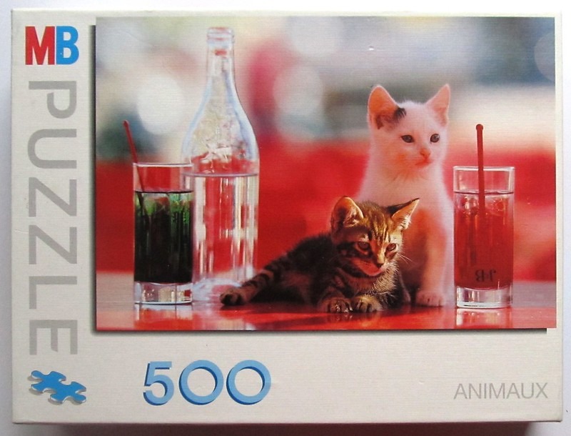 A white kitten and a brown tabby kitten sitting on a red surface next to two tall glasses and a clear glass bottle.