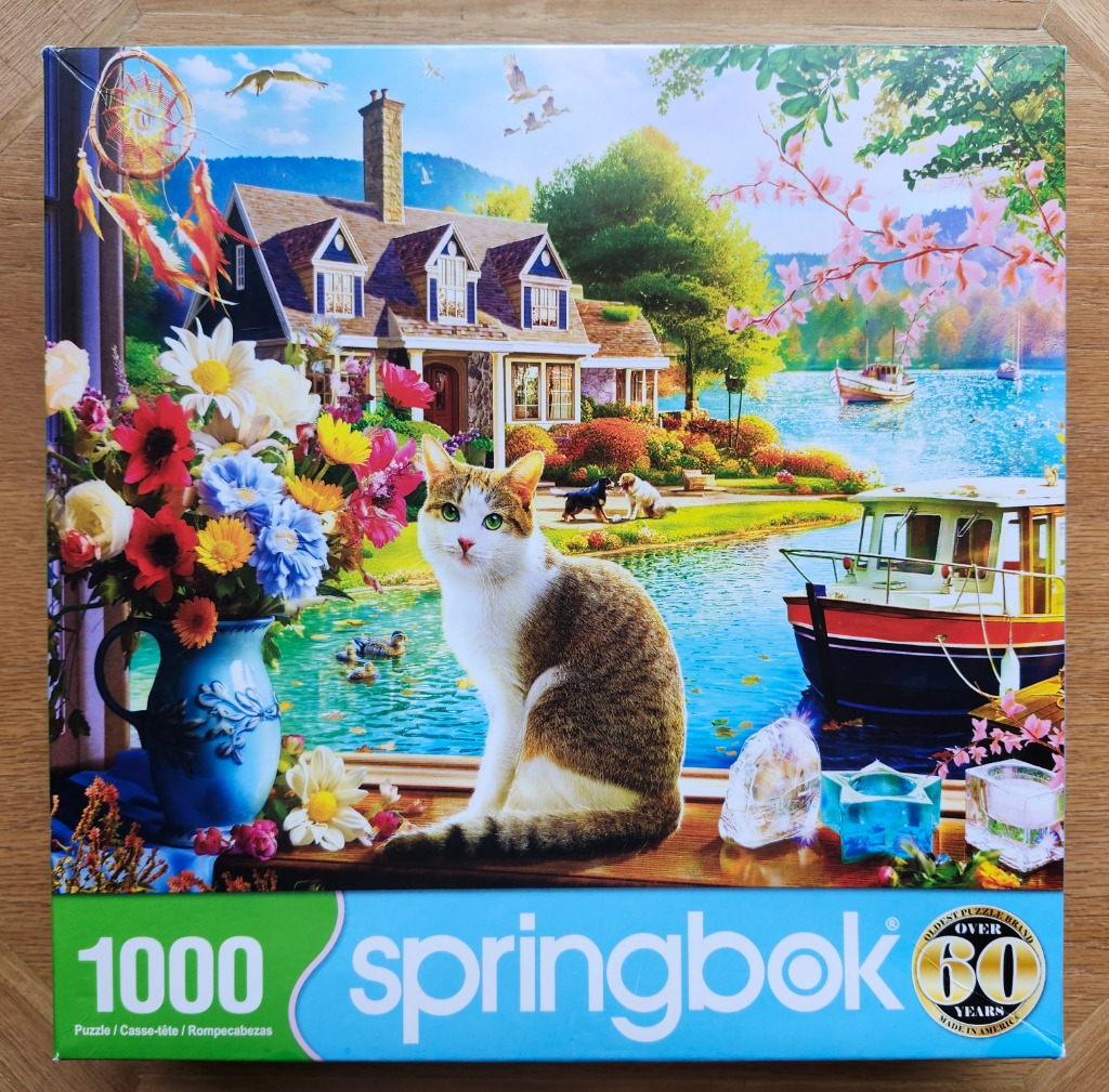 A tabby and white cat sits on a windowsill overlooking a lake scene with a house, a boat, and flowers.