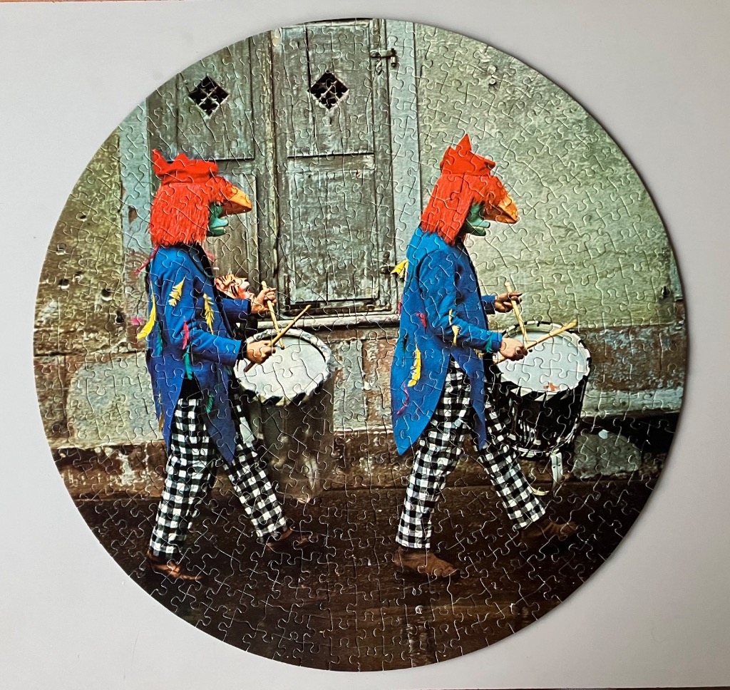 Two figures in red bird masks and vibrant blue coats playing drums in front of an old wall.
