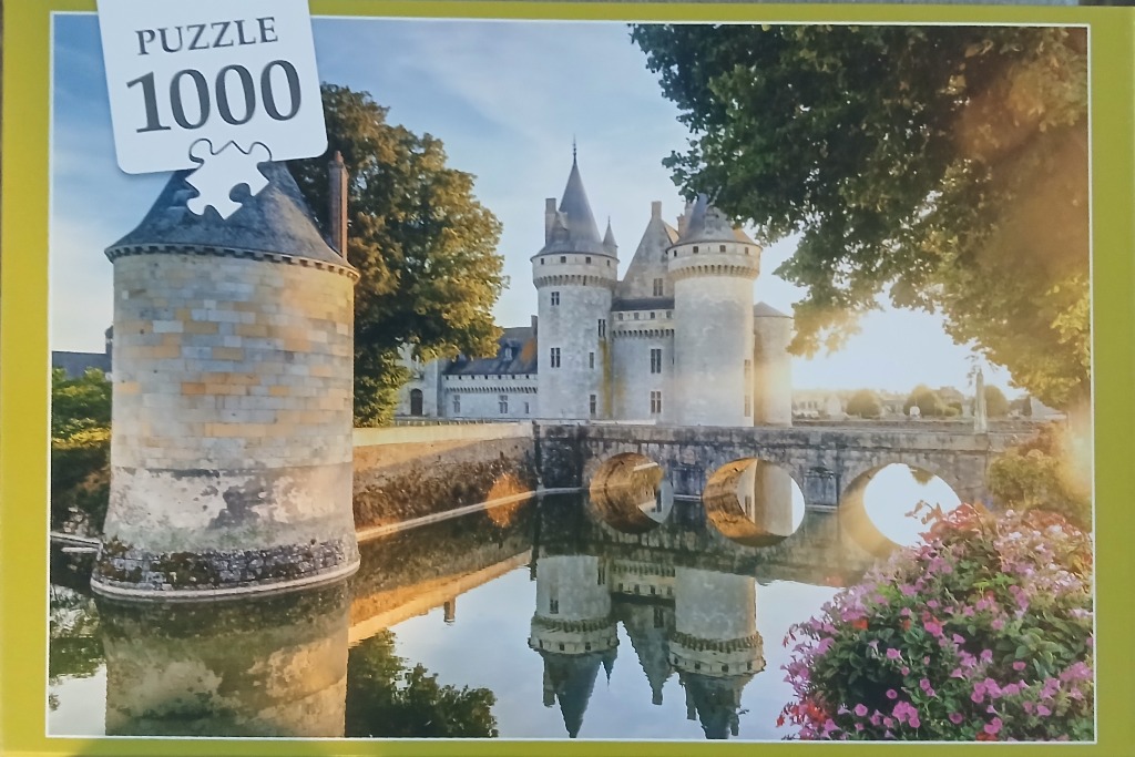 A French-style stone castle with turrets reflected in a moat at sunset, surrounded by trees and flowers.