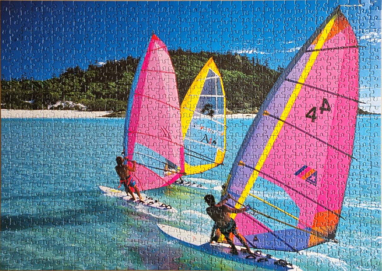 Three windsurfers with colorful sails on bright blue water near a tropical beach.