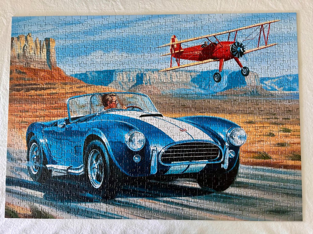 A blue convertible sports car driving through a desert landscape with a red biplane flying above.