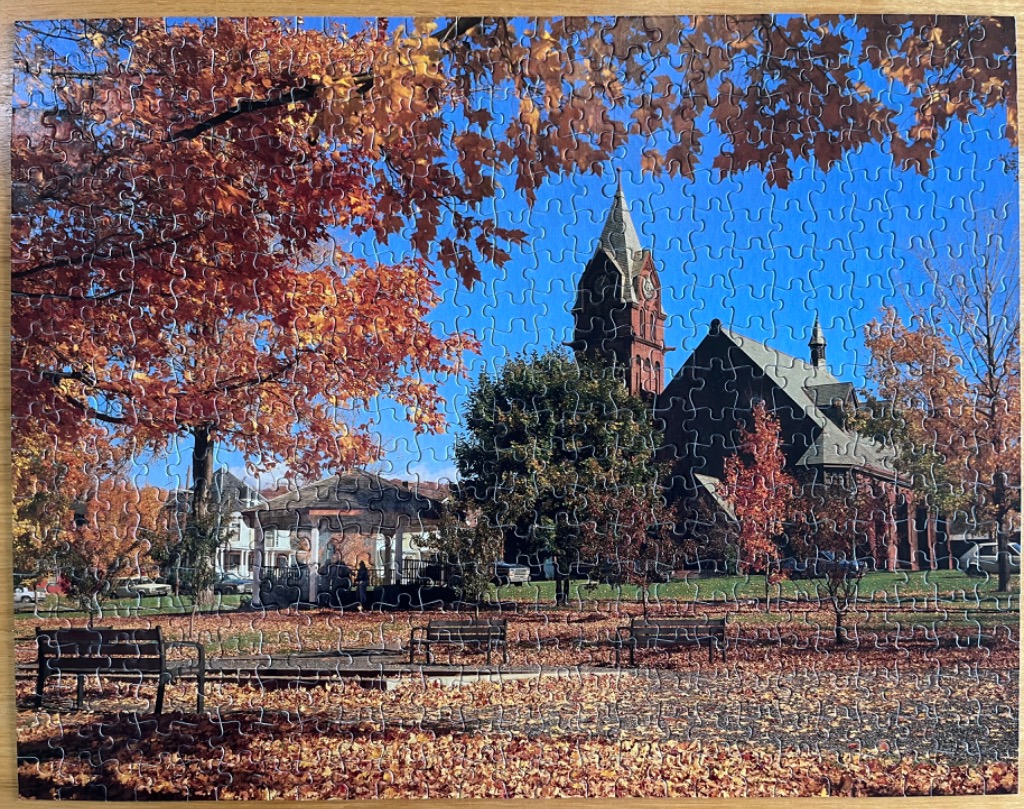A peaceful town square in autumn with a gazebo and a red brick church under a clear blue sky.