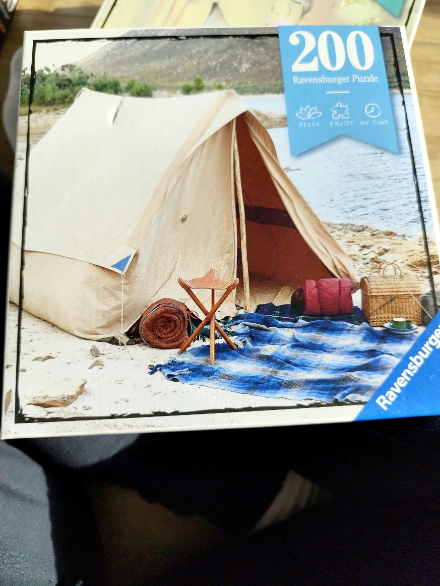 A tan canvas tent pitched on a sandy beach by a lake with a blue picnic blanket, a folding stool, and a wicker basket.