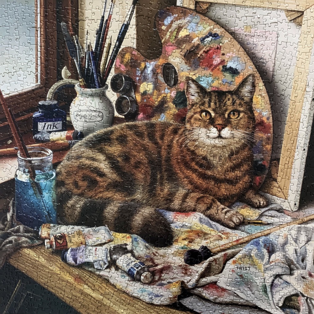 An oil painting of a tabby cat resting on a cluttered artist's table among brushes, paints, and a palette.