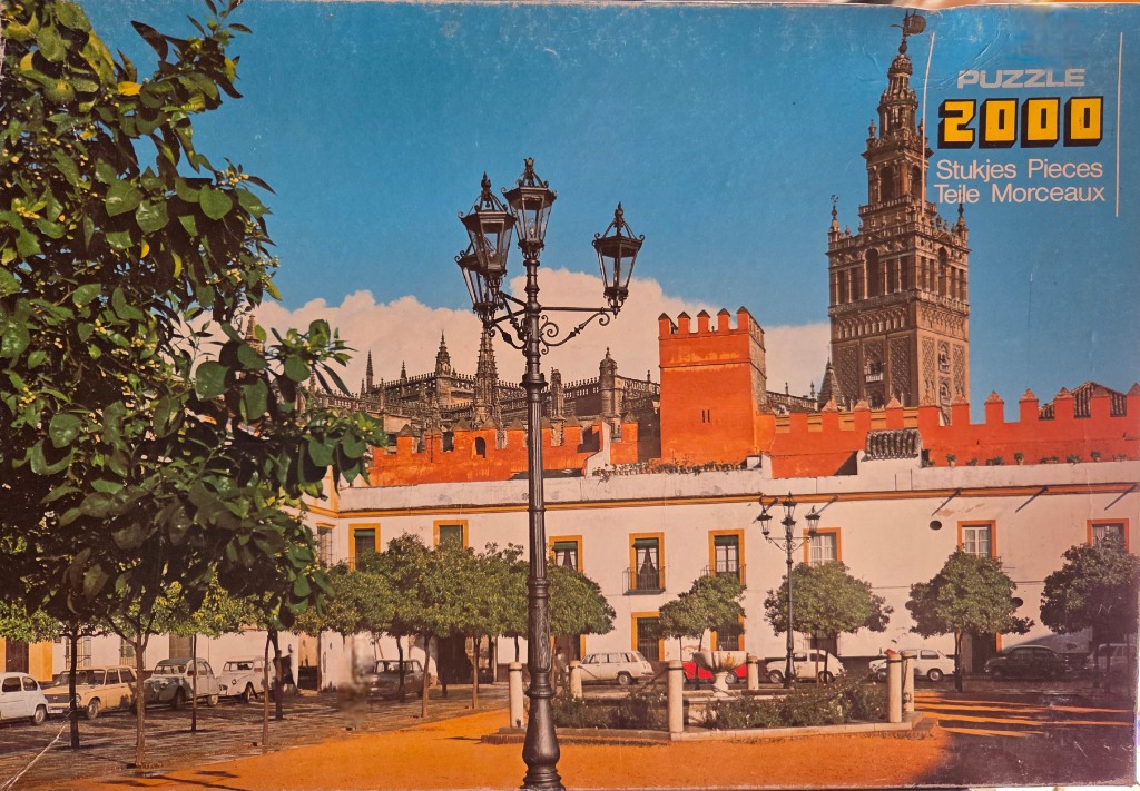 Seville Cathedral and Giralda tower seen from a leafy plaza with a historic black lamp post in the foreground.