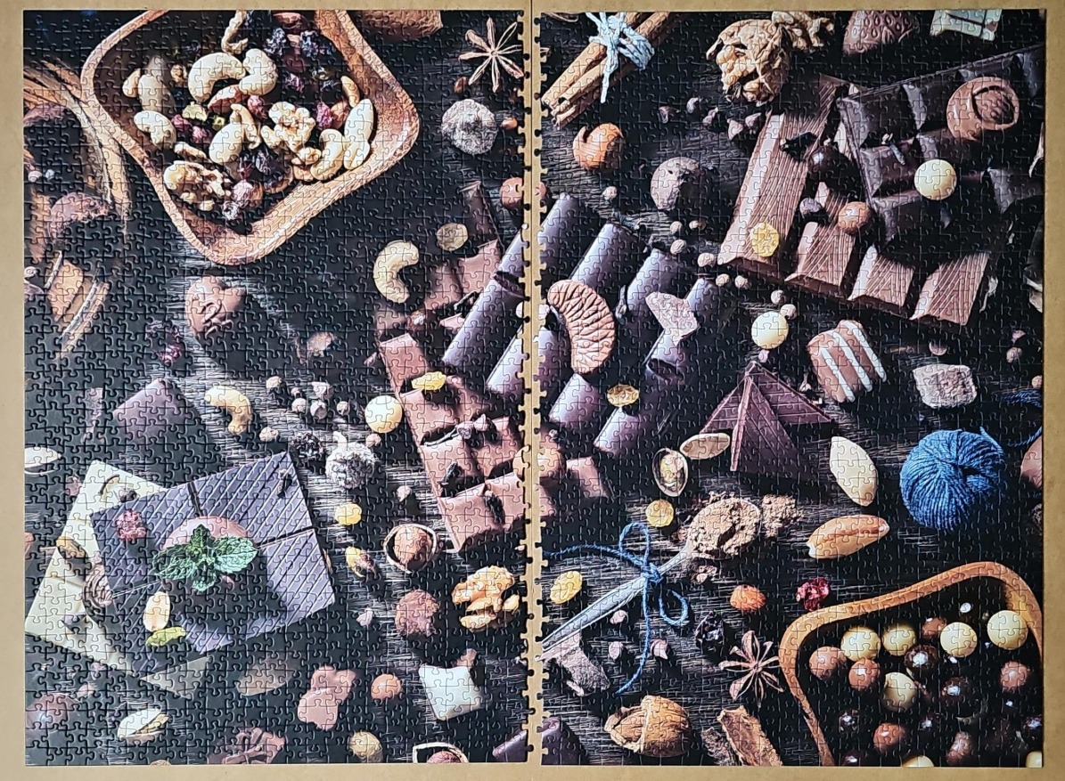 A top-down still life of assorted chocolate bars, nuts, dried fruits, and spices on a dark wooden surface.