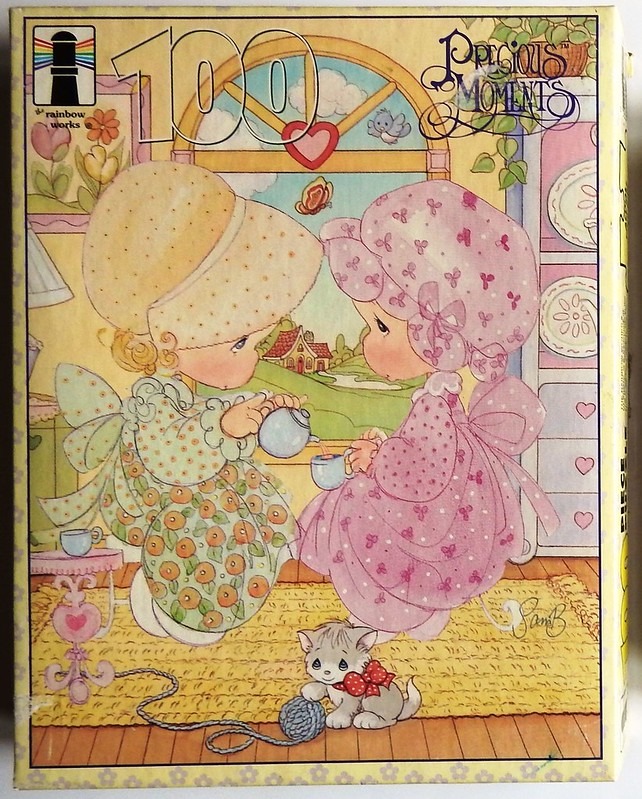 Two young children in Victorian-style dresses and bonnets having a tea party with a kitten on a rug.