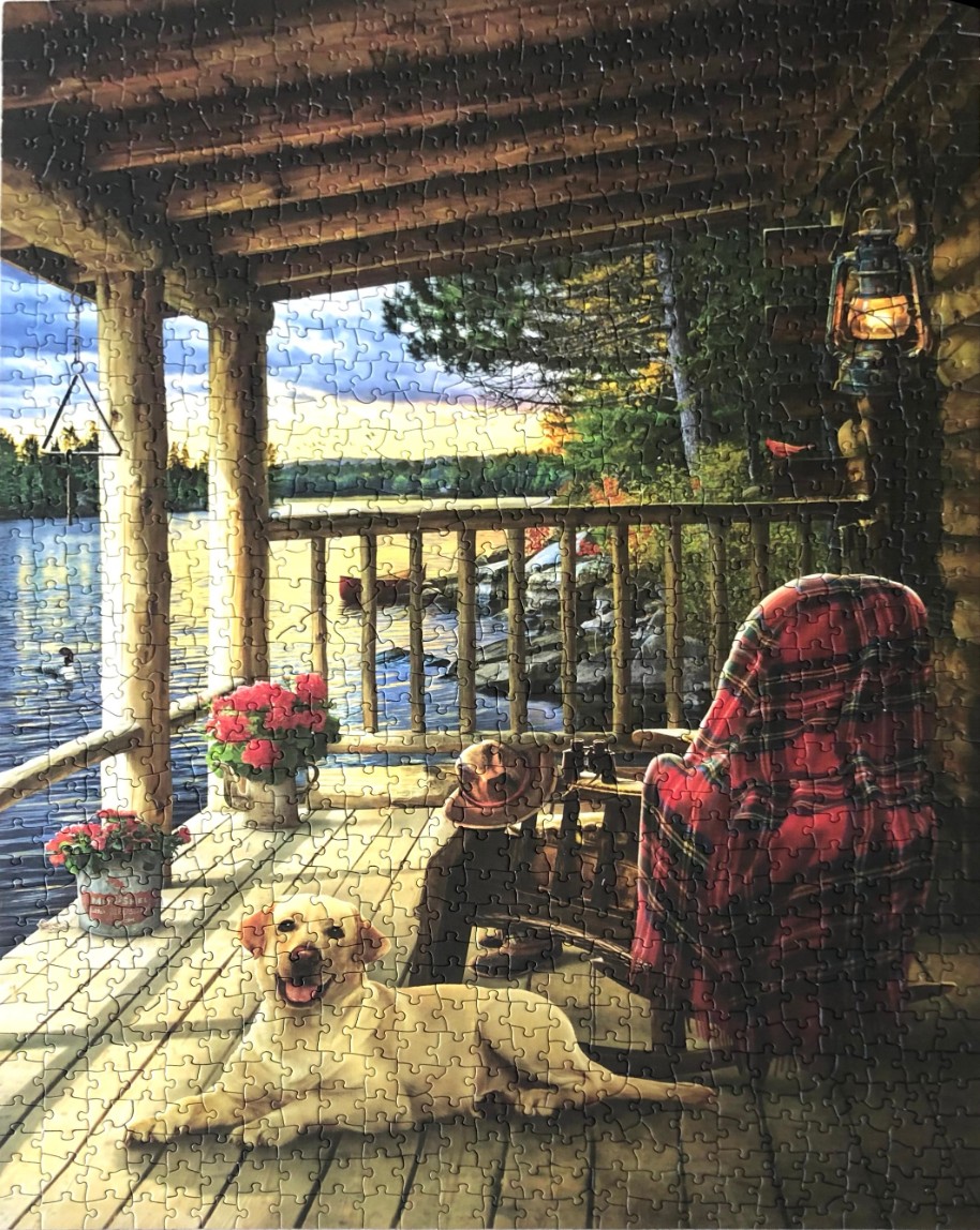 A yellow Labrador lies on a rustic wooden porch overlooking a sunset lake with a plaid chair nearby.