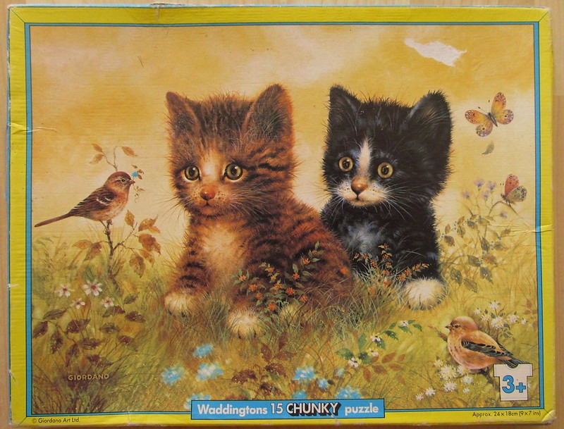 A painting of a tabby kitten and a black kitten sitting in a grassy meadow with birds and butterflies.