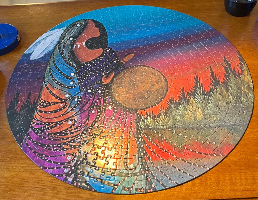 A round illustration of a Native American woman in profile playing a drum against a sunset sky and forest silhouette.
