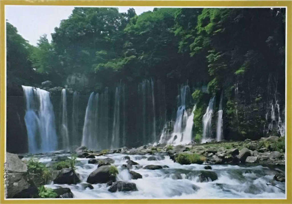 A lush, scenic landscape photograph of Shiraito Falls, featuring multiple thin cascades of water over a rocky cliffside.
