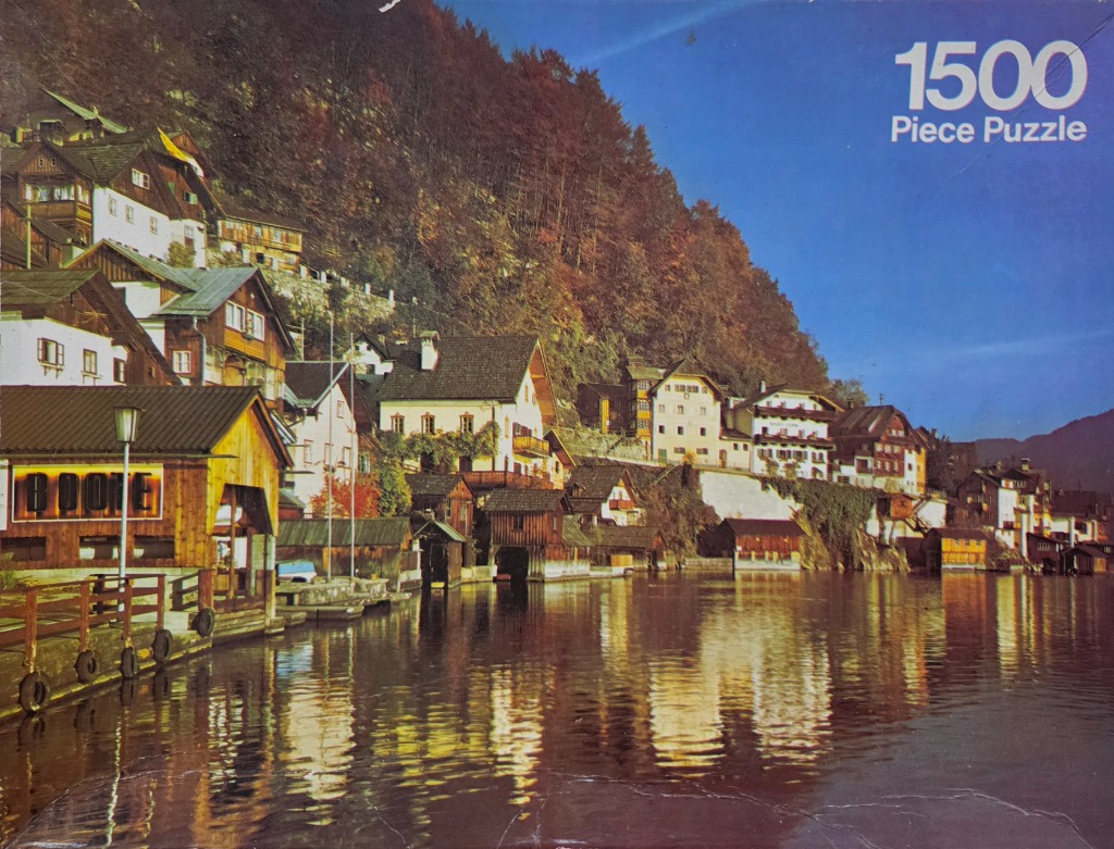 A scenic view of a European village nestled along a lakeshore beneath a steep, forested mountain side.