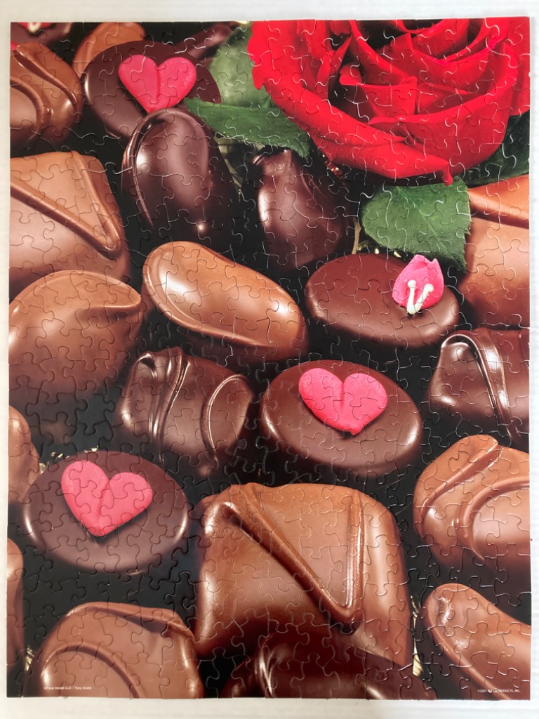 A collection of assorted chocolates accented with a red rose and pink heart decorations.
