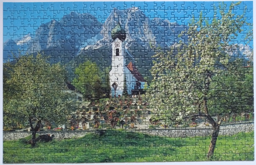 A white church with a green onion dome sits before a massive mountain range in a green, springtime landscape.