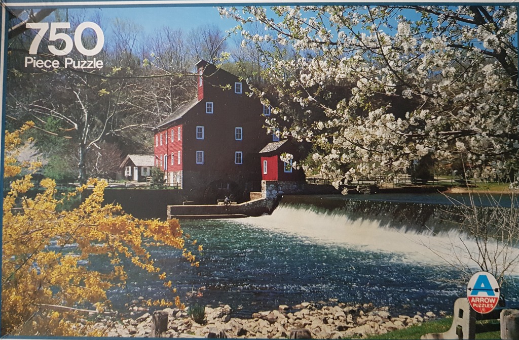 A red mill building sits beside a flowing river with a waterfall, surrounded by blooming yellow and white trees.