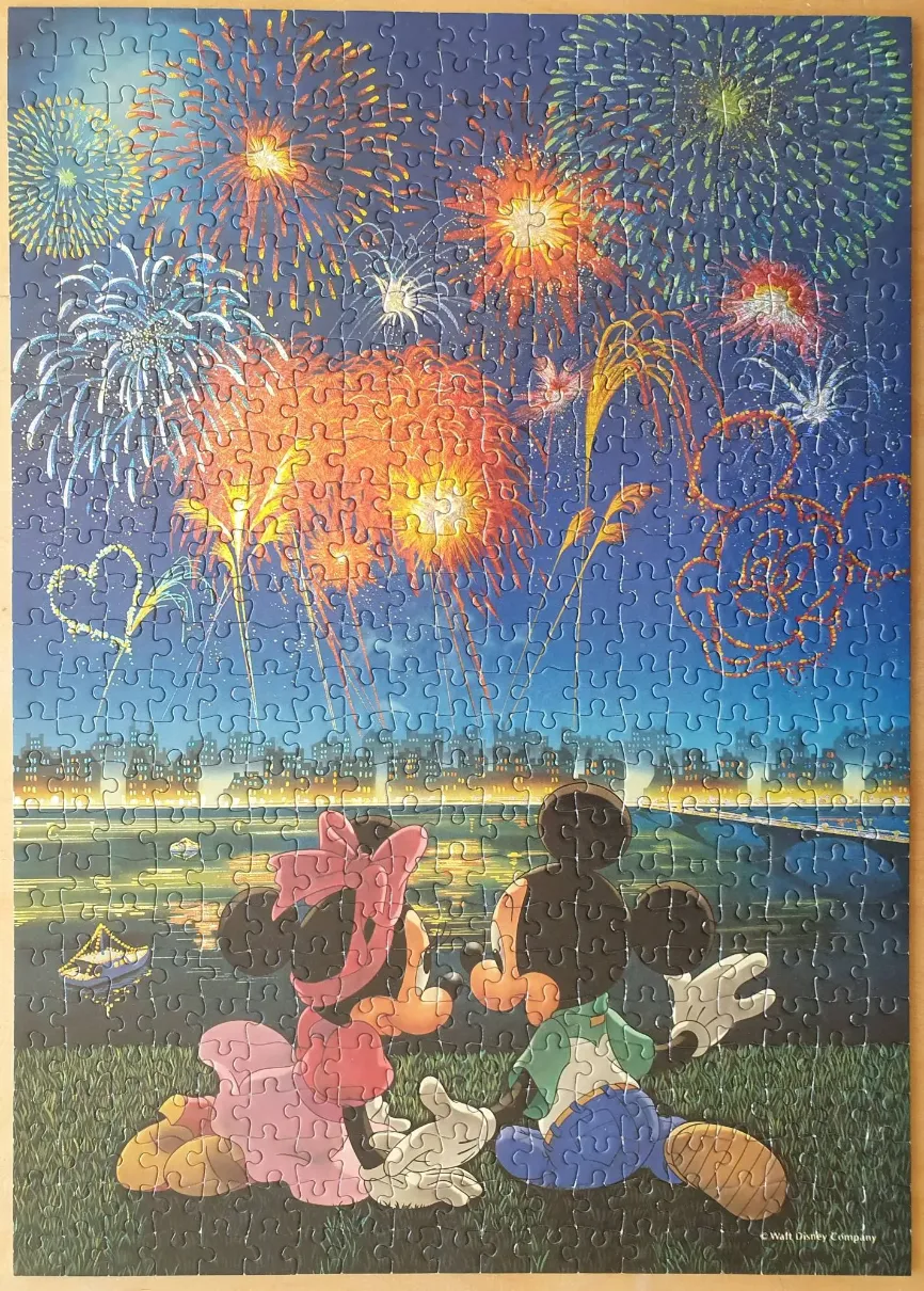 Mickey and Minnie Mouse sitting on a grassy hill at night watching a vibrant fireworks display over a city skyline.