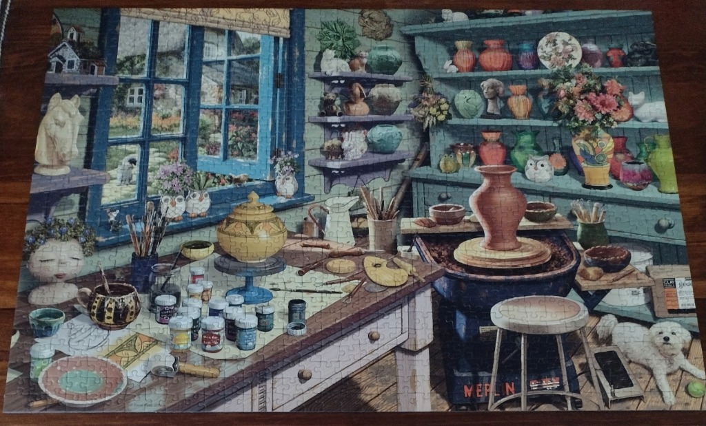 A cozy, rustic pottery studio filled with shelves of ceramic vases, painting tools, a pottery wheel, and a small white dog.
