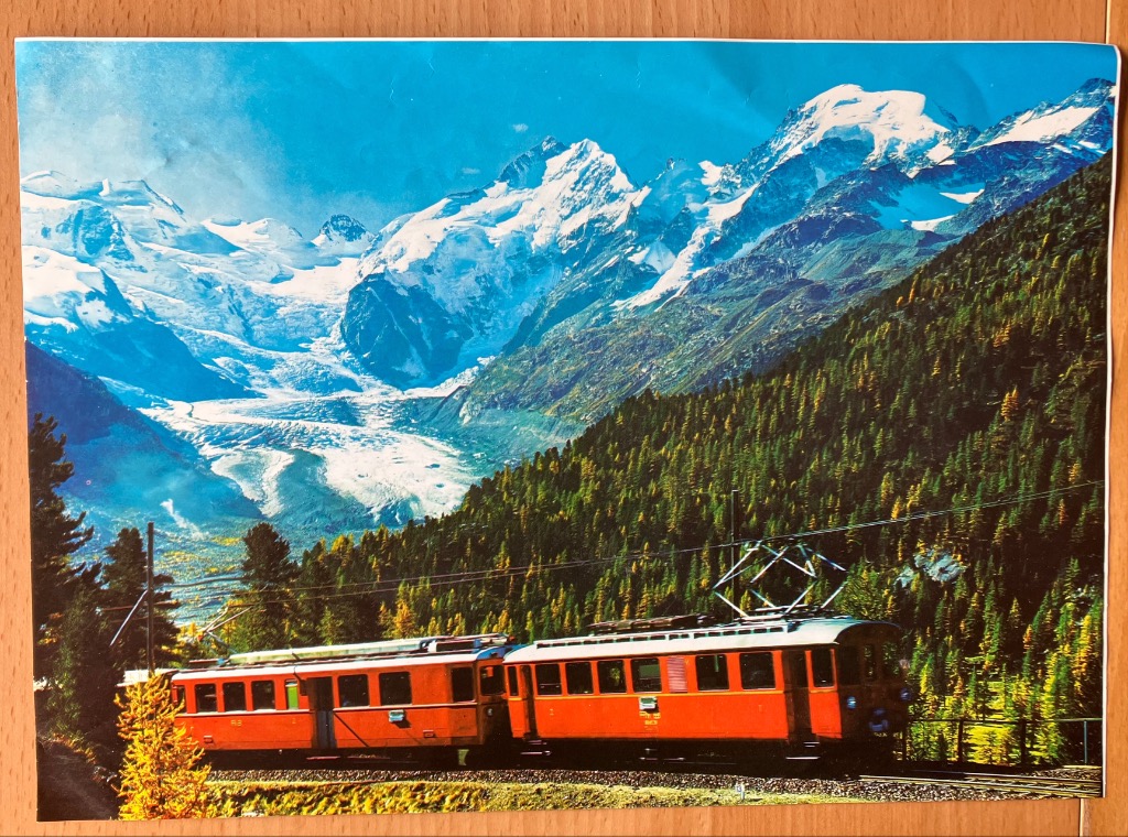 A red train traveling through a pine forest with snow-capped mountains in the background.