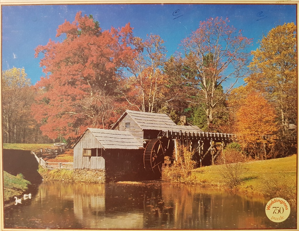 A rustic watermill beside a calm pond surrounded by vibrant autumn trees under a clear blue sky.