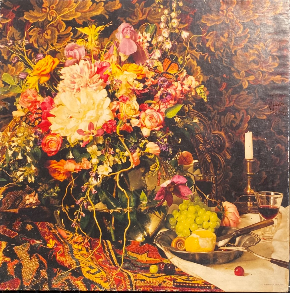 A lush floral still life in a glass bowl on a table with fruit, a candle, and a patterned tapestry background.