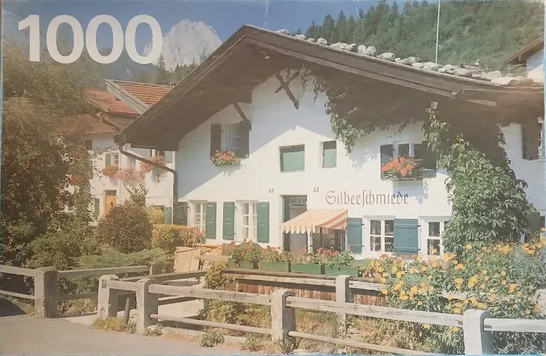 A traditional Alpine house with white stucco walls, green shutters, and overflowing flower boxes, set against mountains.